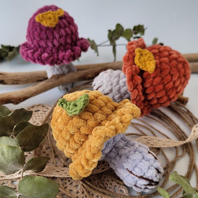 May include: Three crocheted mushroom toys with different coloured caps and stems. The mushrooms are made with fuzzy yarn and have small green leaves on top.