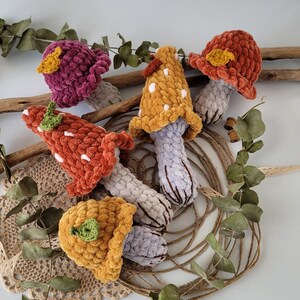 May include: Five crocheted mushroom toys in various colours, including purple, orange, yellow, and grey. The mushrooms have white polka dots and green leaves.