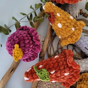 May include: Four crocheted mushroom toys in different colours and patterns. The mushrooms are made with fuzzy yarn and have white polka dots. One mushroom has a yellow leaf, another has a green leaf, and the other two have white spots.
