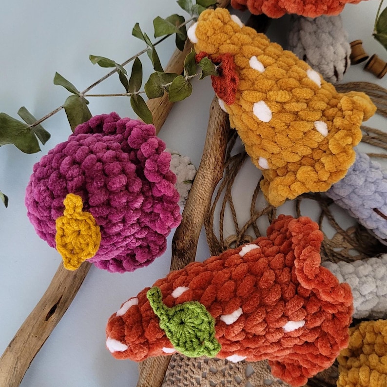 May include: Four crocheted mushroom toys in different colours and patterns. The mushrooms are made with fuzzy yarn and have white polka dots. One mushroom has a yellow leaf, another has a green leaf, and the other two have white spots.