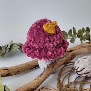May include: A crocheted mushroom toy with a pink cap and a yellow top. The mushroom is sitting on a wooden branch.