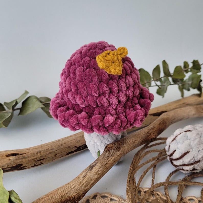 May include: A crocheted mushroom toy with a pink cap and a yellow top. The mushroom is sitting on a wooden branch.