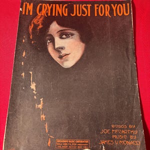 May include: A vintage sheet music cover with a black background and a woman's face in the center. The title of the song is "I'm Crying Just For You" and the words and music are credited to Joe McCarthy and James V. Monaco. The cover also includes the Broadway Music Corporation logo.