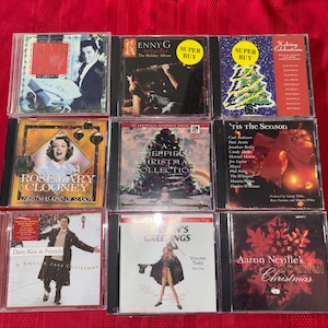 May include: Nine CD cases featuring various artists and holiday albums. Titles include "Enny G The Holiday Album", "Rosemary Clooney Christmas Kind of Season", and "Aaron Neville's Soulful Christmas". The CDs are displayed on a red surface.