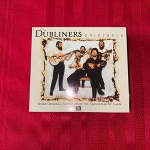 Puede incluir: Una caja de CD con el título "The Dubliners Originals" y una imagen de la banda tocando instrumentos. La caja tiene un borde color crema con el texto "Three Original Albums from the Transatlantic Label". El fondo es una tela roja.