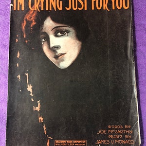 May include: A vintage sheet music cover with a black background and a woman with dark hair and a tear on her cheek. The title of the song is "I'm Crying Just For You" and the songwriters are Joe McCarthy and James V. Monaco. The sheet music is published by Broadway Music Corporation.