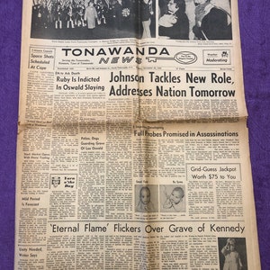 May include: A black and white newspaper front page with the headline "Johnson Tackles New Role, Addresses Nation Tomorrow". The article discusses the assassination of President John F. Kennedy and the subsequent investigation. The newspaper is dated November 24, 1963.