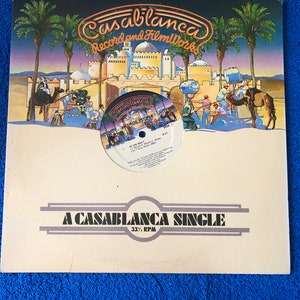 Może przedstawiać: Biała okładka płyty winylowej z obrazkiem przedstawiającym scenę pustynną z wielbłądem i ludźmi. Tekst "Casablanca Record and FilmWorks" znajduje się w górnej części okładki. Tekst "A Casablanca Single 33 1/3 RPM" znajduje się w dolnej części okładki.