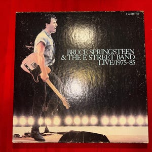 Puede incluir: Una caja negra de Bruce Springsteen & The E Street Band Live/1975-85 que contiene 3 casetes. La portada presenta una fotografía de un músico con camisa clara y pantalones oscuros tocando una guitarra eléctrica clara en un escenario iluminado.