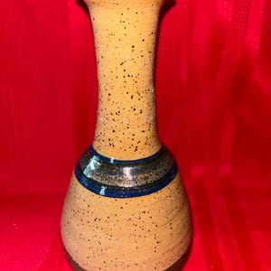 May include: A speckled, handcrafted ceramic vase with a flared top and a rounded base. The vase features a warm yellow-brown color with dark speckles and blue and brown bands around the neck. The rim is a dark blue.