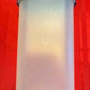 Op de afbeelding: Een hoge, doorschijnende plastic container met een donkerblauw deksel. De container heeft een rechthoekige vorm met afgeronde hoeken. De achtergrond is effen rood.