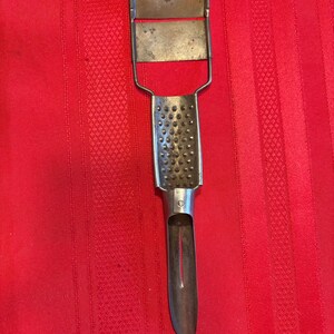 Op de afbeelding: Vintage metalen keukengereedschap met een mes en een rasp. Het gereedschap heeft een handvat en een mes aan de onderkant. Het rasp gedeelte heeft kleine, ronde gaatjes. Het gereedschap is van metaal en wordt waarschijnlijk gebruikt voor voedselbereiding.