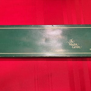 May include: A rectangular green box with a white border and the text "The Master Carver" and "Frontier Forge" in white script. The box is set against a red background.