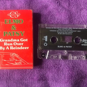 May include: A red cassette tape with the title "Grandma Got Run Over By A Reindeer" by Elmo & Patsy. The cassette tape is in a clear plastic case with a black label. The label includes the song title, artist names, and copyright information. The cassette tape is next to a red cassette single cover with the same title and artist names.
