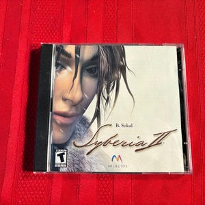 May include: A CD case for the video game Syberia II, featuring a woman with brown hair and a fur-lined jacket on the cover. The title of the game is written in cursive with the text "B. Sokal" above it. The Microïds logo is below the title.