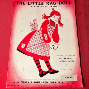 May include: A vintage sheet music cover featuring a red rag doll with black and white striped stockings and red and white checkered dress. The title of the song is "The Little Rag Doll with the Shoe Button Eyes". The sheet music is by Milton Pascal and Gerald Marks. The price is 60 cents.