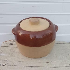 May include: A round ceramic crock with a lid, featuring a two-tone design with a glossy dark brown glaze on the upper portion and a matte light beige finish on the lower section. The pot includes two small side handles and a central knob on the lid.