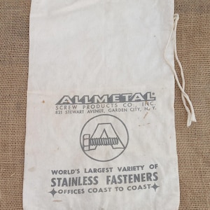 May include: A white canvas bag with a drawstring closure. The bag has a black printed logo that reads "ALLMETAL SCREW PRODUCTS CO., INC. 821 STEWART AVENUE, GARDEN CITY, NY." and "WORLD'S LARGEST VARIETY OF STAINLESS FASTENERS OFFICES COAST TO COAST".