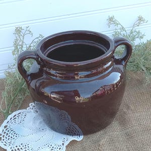 May include: A large, dark brown ceramic crock with two handles. The crock has a smooth, glossy finish and is sitting on a burlap surface.
