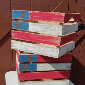 May include: Two wooden crates stacked on top of each other. Each crate is painted with a red, white, and blue design, featuring a star on the blue section. The crates have a rustic, distressed finish, suggesting a vintage or handmade aesthetic.