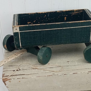 May include: A vintage wooden toy wagon painted dark green with white accents. The wagon has four round wheels and a pull string. The paint shows signs of wear, giving it a rustic appearance. A classic toy for children.