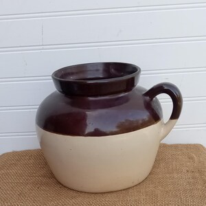 May include: A round ceramic crock featuring a two-tone design with a dark brown glazed upper section and a smooth cream-colored lower body. The vessel includes a wide, thick rim and a single curved handle attached to the side of the pot.