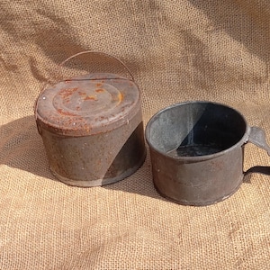 May include: Two antique metal containers. One is a round, lidded bucket with a handle, showing rust. The other is a cup-shaped container with a handle. Both have a weathered, aged appearance, suggesting they are vintage items.