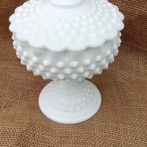 May include: A white hobnail candy dish with a lid, set on a pedestal base. The dish has a textured surface with small, rounded bumps and a scalloped edge. The lid is topped with a small, decorative finial.