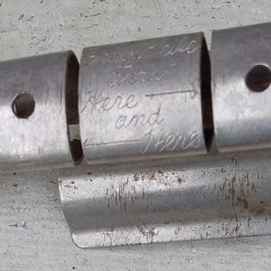 May include: A metallic, cylindrical tool featuring three distinct segments. The central segment is engraved with the text "Insert Here and Here" accompanied by an arrow. The metal surface shows signs of wear, including minor scratches and oxidation.