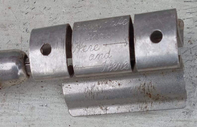 May include: A metallic, cylindrical tool featuring three distinct segments. The central segment is engraved with the text "Insert Here and Here" accompanied by an arrow. The metal surface shows signs of wear, including minor scratches and oxidation.