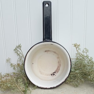 May include: A vintage black and white enamel saucepan with a long handle. The interior is off-white with dark staining. The handle has a hole for hanging. The pan is set against a white background with sprigs of greenery.