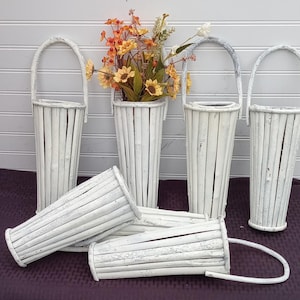 May include: A collection of white, twig-style hanging flower baskets, some upright and some lying down. One basket holds a bouquet of yellow and orange flowers. The baskets have arched handles and a rustic, weathered appearance.