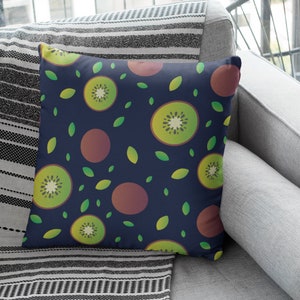 Puede incluir: Una almohada azul marino con un patrón repetitivo de rodajas de kiwi y hojas verdes. Las rodajas de kiwi son de color verde claro con un centro marrón. La almohada está sentada en un sofá a rayas.