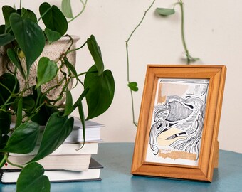May include: A wooden framed artwork with a black and white abstract design, displayed on a teal table. A potted plant with green leaves and trailing vines is next to the artwork. Books are stacked near the plant.