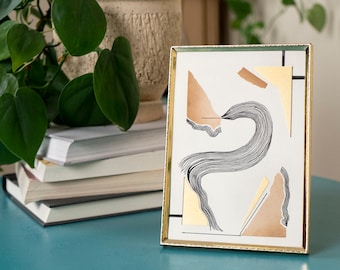 May include: Abstract art print in a gold frame. The artwork features geometric shapes in gold and tan, with a central black line drawing. The frame is rectangular and sits on a teal surface, with a plant and books in the background.
