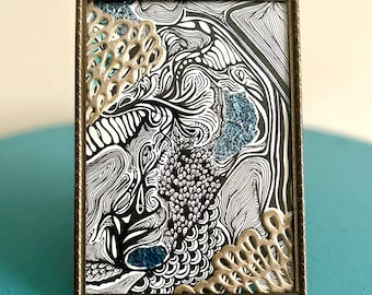 Puede incluir: Obra de arte abstracta en un marco rectangular de bronce. La obra presenta dibujos lineales en blanco y negro con detalles en azul y adornos decorativos plateados. La obra se exhibe sobre una superficie turquesa.