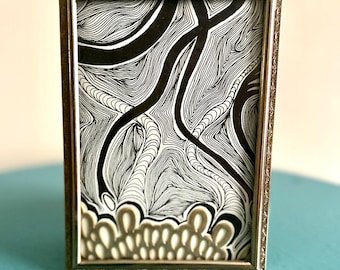Puede incluir: Una obra de arte abstracta enmarcada. La obra presenta un diseño en blanco y negro con líneas y formas fluidas. El marco es de color plateado metálico con detalles ornamentados. La obra de arte se exhibe sobre una superficie azul.