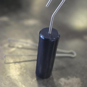 May include: A cylindrical dark blue weight with a smooth finish, featuring a silver-colored metal hook extending from the top center. The object is positioned upright, with two additional thin metal wire hooks visible in the background.