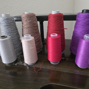 May include: A collection of eight sewing thread cones arranged in two rows. The back row features larger cones in light gray, beige, red, and purple. The front row contains smaller cones in light gray, pale pink, bright red, and vibrant purple.
