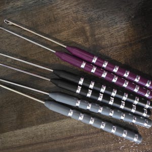May include: A set of six latch hook tools with long metal shafts and hooked tips. The handles are cylindrical with a series of silver-colored rings and come in three colors: two purple, two black, and two gray, arranged in a diagonal pattern.