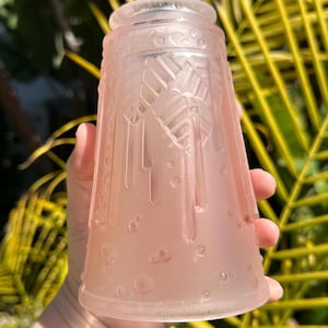 May include: A pink glass lampshade with an Art Deco design. The shade is a soft pink color and features etched geometric patterns and vertical lines. The lampshade has a flared shape and a rounded top. The background is blurred with green and yellow foliage.