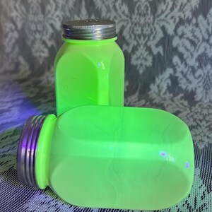 Könnte beinhalten: Zwei neongrüne Glasbehälter mit silbernen Deckeln. Ein Behälter steht aufrecht, der andere liegt auf der Seite. Die Behälter haben eine quadratische Form mit abgerundeten Kanten. Der Hintergrund ist ein gemustertes weißes und graues Gewebe.