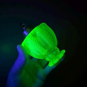 Può includere: Una coppa di vetro verde brillante con un motivo floreale, tenuta in mano. La coppa ha uno stelo e una base svasata. Il vetro si illumina su uno sfondo scuro.