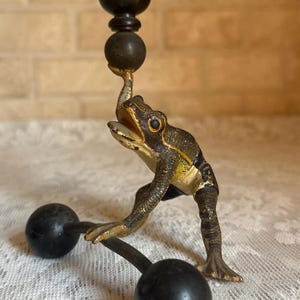 May include: A whimsical, antique-style candle holder featuring a frog figurine. The frog is bronze-colored with gold accents, supporting a black candle cup. Two black spheres are attached to the base, adding to the unique design.