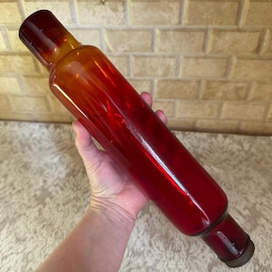 May include: A red glass rolling pin with a gradient of orange at the top. The rolling pin is cylindrical with a metal cap on one end. The object is held in a hand.