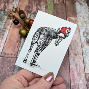 May include: A black and white illustration of an okapi wearing a red Santa hat on a white card. The okapi has zebra-like stripes on its hindquarters. The card is held by a person with a wooden background.