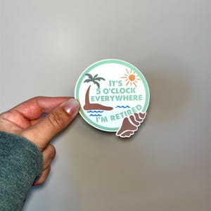 May include: A round sticker with a beach theme. The sticker has a green border and features a palm tree, sun, and waves. Text on the sticker reads "It's 5 o'clock everywhere, I'm retired."