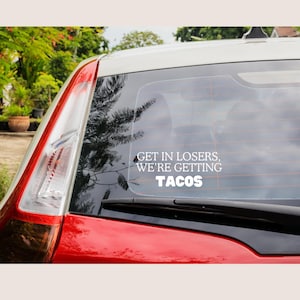 May include: A white decal on a car's rear window with the text "GET IN LOSERS, WE'RE GETTING TACOS." The car is red and white, with a view of trees and sky visible through the window.