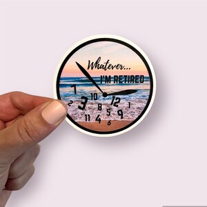 May include: Round sticker with a beach scene and the words "Whatever... I'M RETIRED" in black. The sticker has a black border and the clock hands point to 12:00. The background shows a sunset over the ocean.
