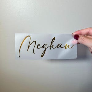 May include: A white rectangular decal with the name "Meghan" in elegant gold script. The decal is held by a hand with painted nails against a plain white wall. The decal is likely for personalization.